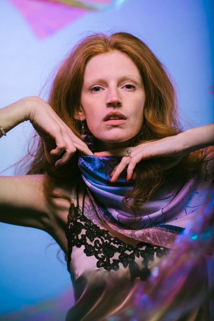 A woman with long, red hair wearing a silky, patterned top with black lace detail, posing with her hands near her face against a vibrant, colorful background.