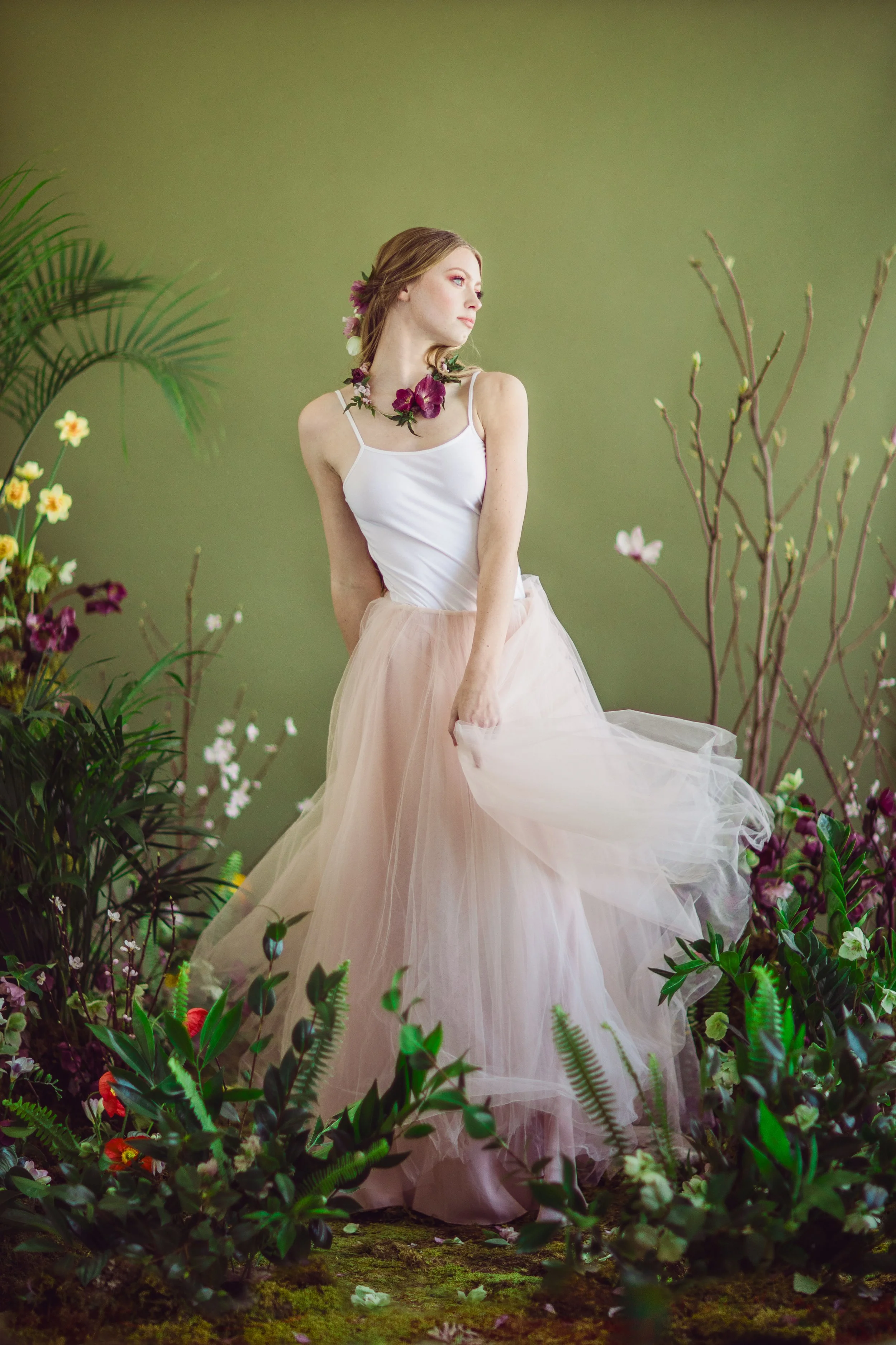 A woman in a white tank top and a flowing pale pink tulle skirt stands amidst lush green plants and flowers, looking to the side with a thoughtful expression. She has a floral necklace and a similar flower in her hair, against a plain green backgroun