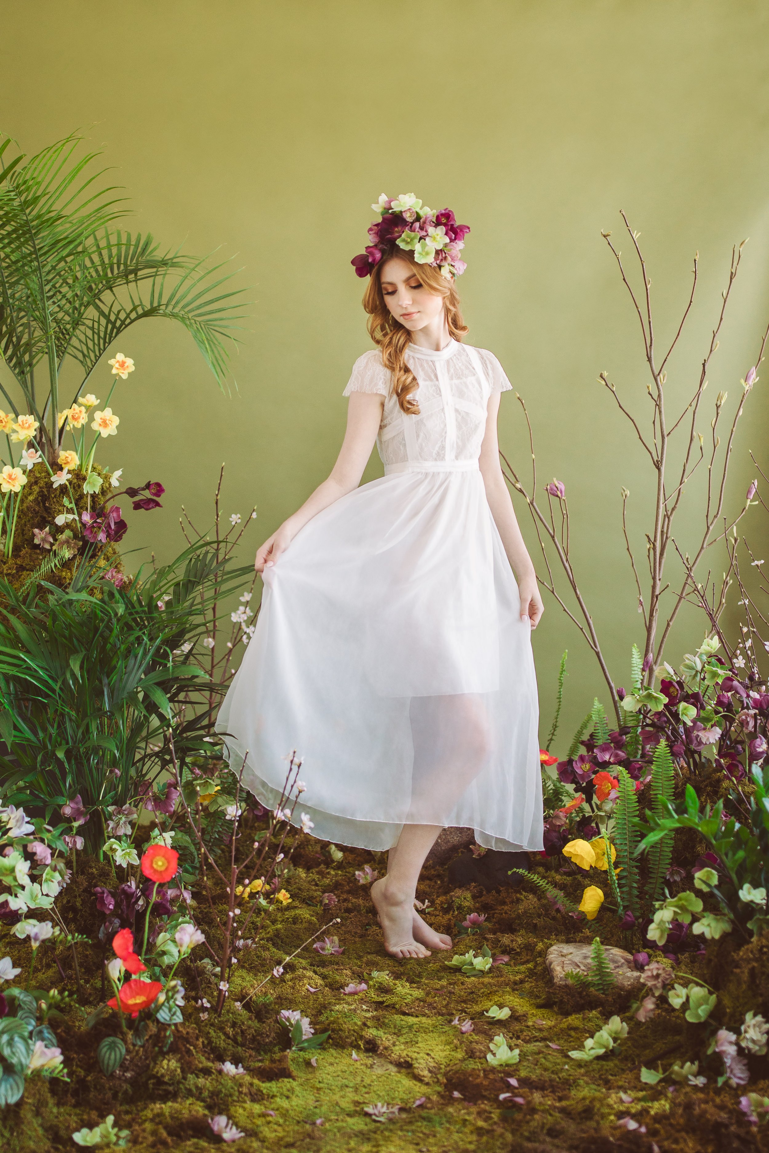 Young woman dressed in a white gown with a floral crown standing among lush green plants and colorful flowers on a mossy ground, with a green background.