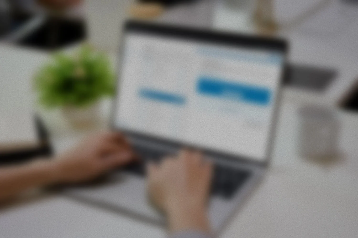 Blurred image of person typing on laptop, with a plant on the desk