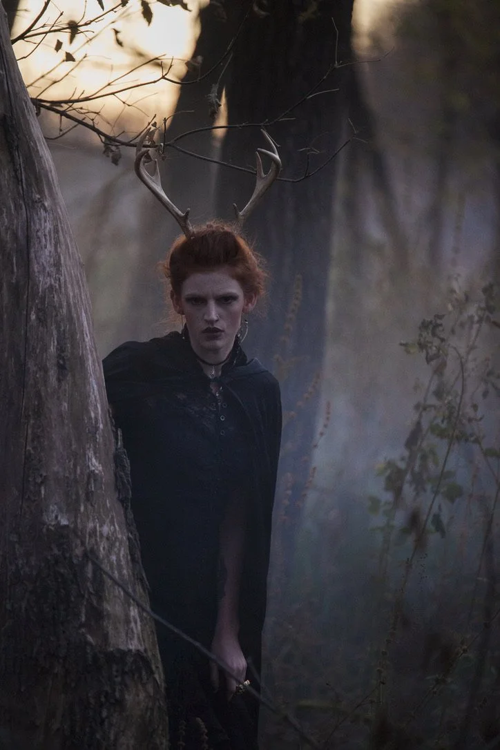 A woman with red hair wearing a black cloak and antler headpiece standing behind a tree in a misty forest at dusk.
