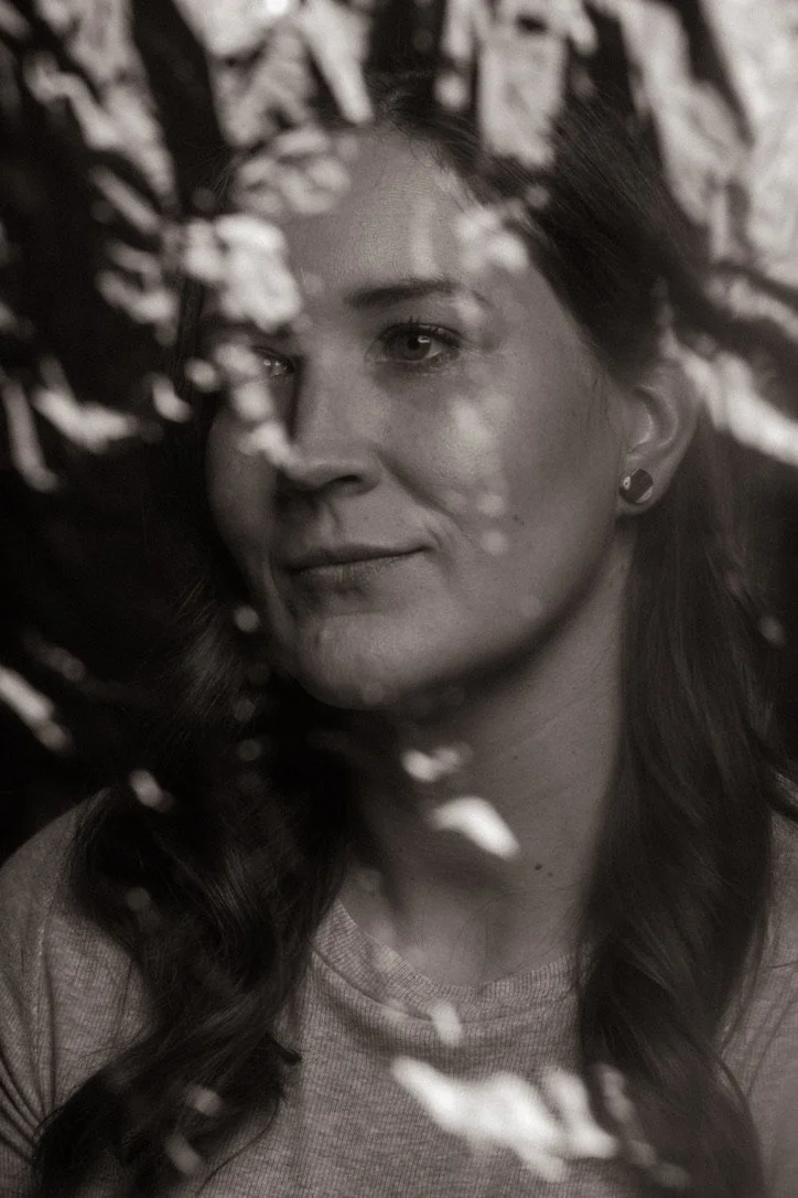 A black and white photo of a woman with long hair, looking thoughtfully through leaves or branches.