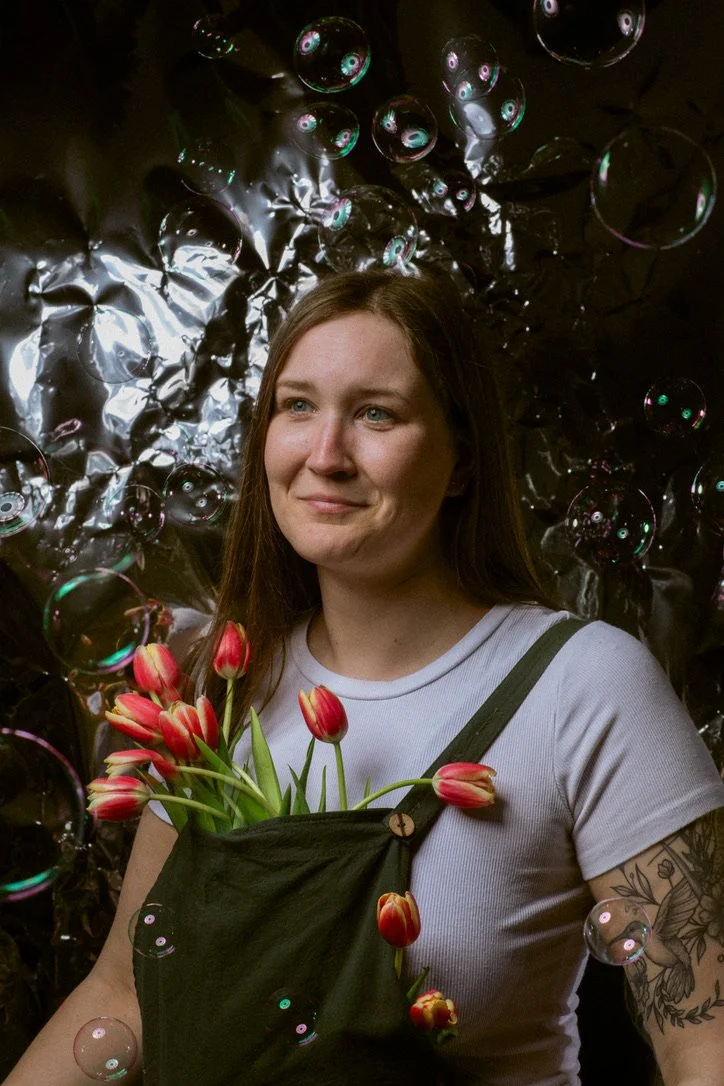 A woman with long brown hair and a tattoo on her arm holding a bunch of red and yellow tulips. She is wearing a white t-shirt and a black apron. The background is decorated with shiny metallic and black material, with floating soap bubbles.