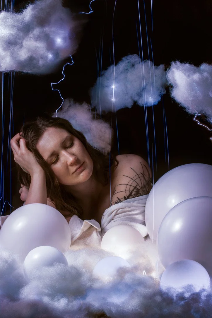 Woman surrounded by artificial clouds and white balloons, with closed eyes and a dreamy expression.