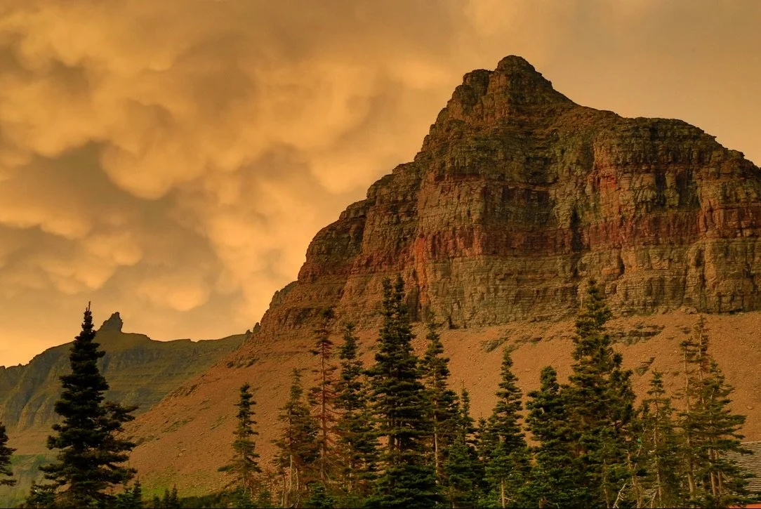 A rugged mountain with layered rocky terrain surrounded by tall evergreen trees, under a sky filled with orange-tinted clouds at sunset or sunrise.