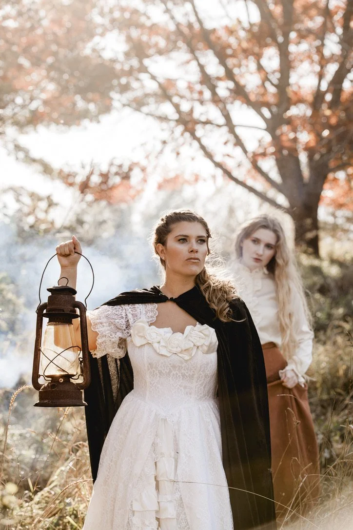 Two women in vintage clothing stand outdoors during autumn, one holding an old lantern, with colorful fall trees and soft sunlight in the background.