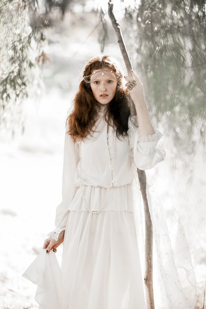 A young woman with long, wavy red hair dressed in a flowing white vintage-style dress, holding a stick, standing outdoors in a wooded area with sunlight filtering through trees.