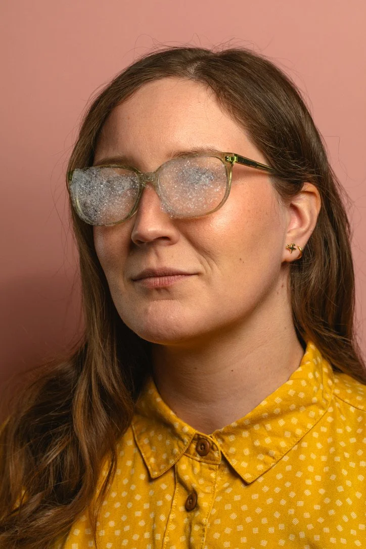 Person wearing glasses with fogged-up lenses, wearing a yellow polka dot shirt, against a peach background.