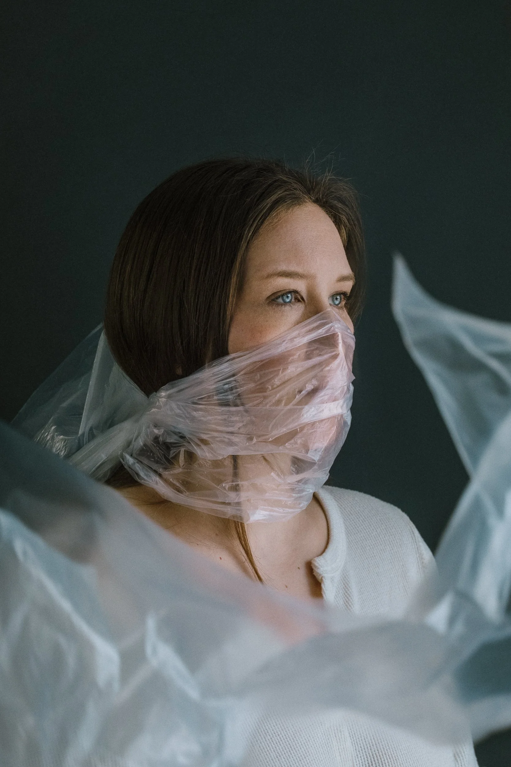 A woman with blue eyes and brown hair, wearing a white shirt, has her face partially covered with a clear plastic bag wrapped around her head and mouth, against a dark background.