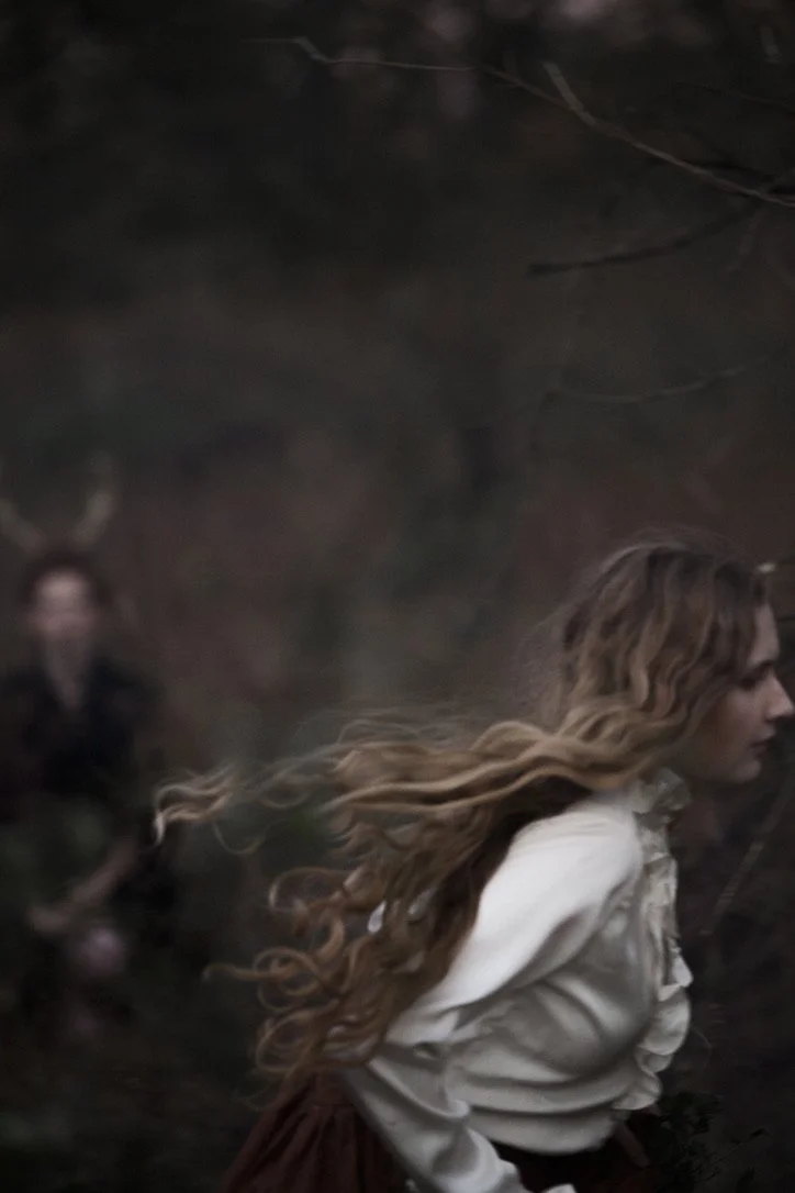 A woman with long, wavy blonde hair running in a dark, forested area with a mysterious figure with antlers in the background.