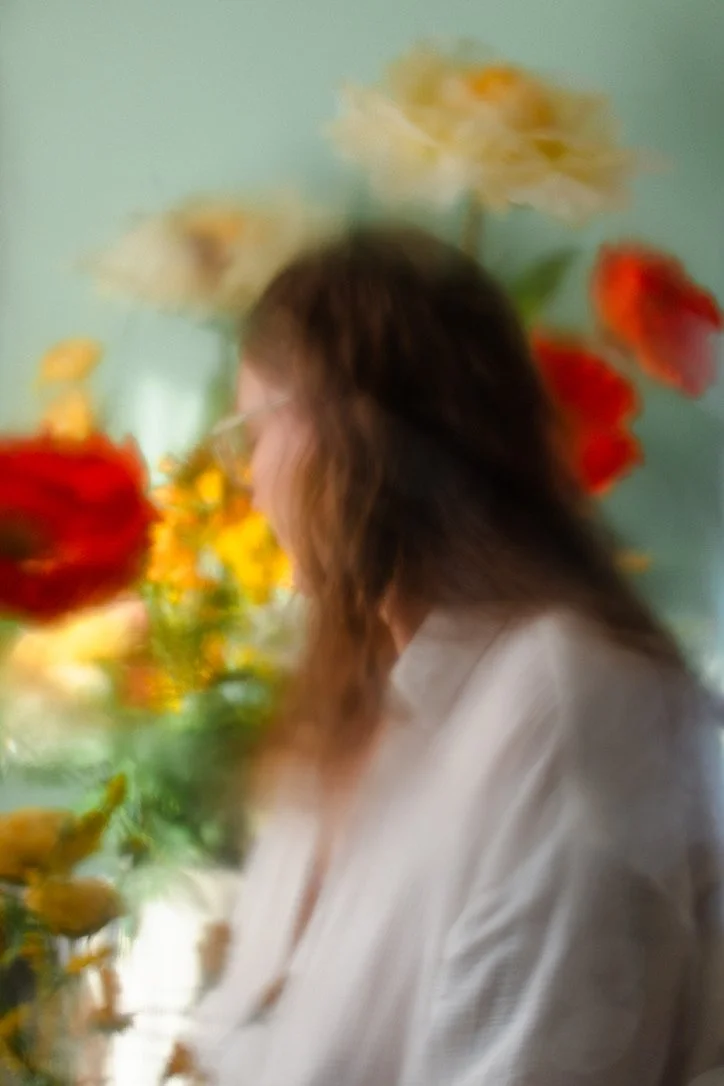 Blurry image of a woman with long brown hair and glasses, standing amidst a colorful background of yellow, red, and white flowers.