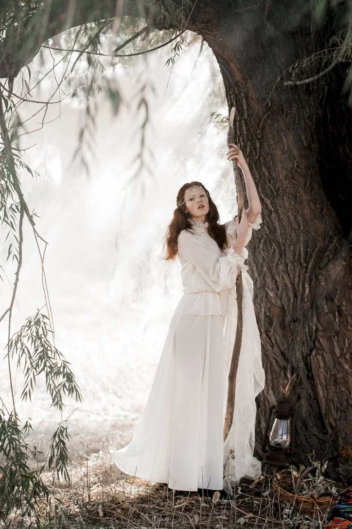 A young woman in a white flowing dress standing next to a large tree, holding a long stick, with a lantern on the ground nearby, in a dreamy, forest setting.