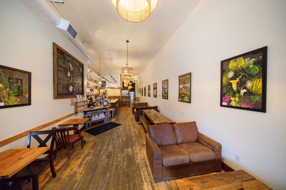 Interior of a cozy café with wooden floor, brown leather couches, framed botanical artwork on white walls, and a counter in the background.