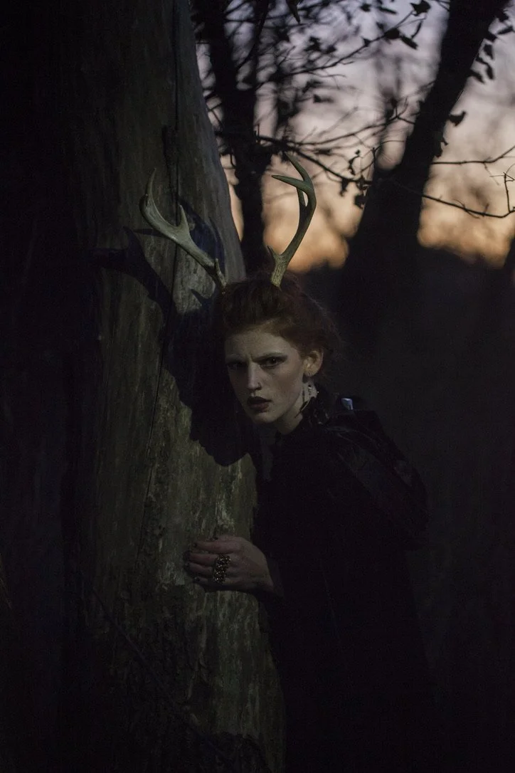 A woman with red hair and antlers, dressed in dark clothing, leaning against a tree at dusk or dawn in a forested area with a sunset or sunrise sky in the background.