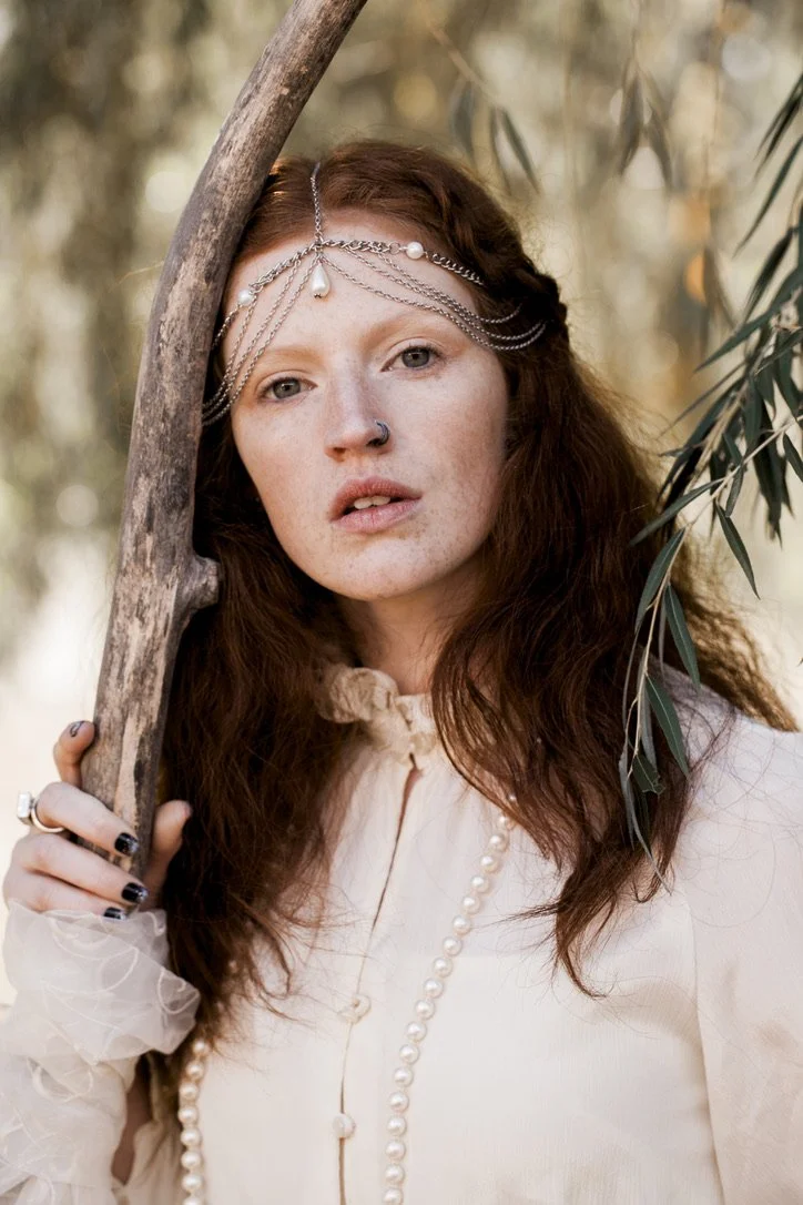 A young woman with long red hair, wearing a pearl headpiece and a cream-colored blouse, leaning against a tree in a natural outdoor setting.