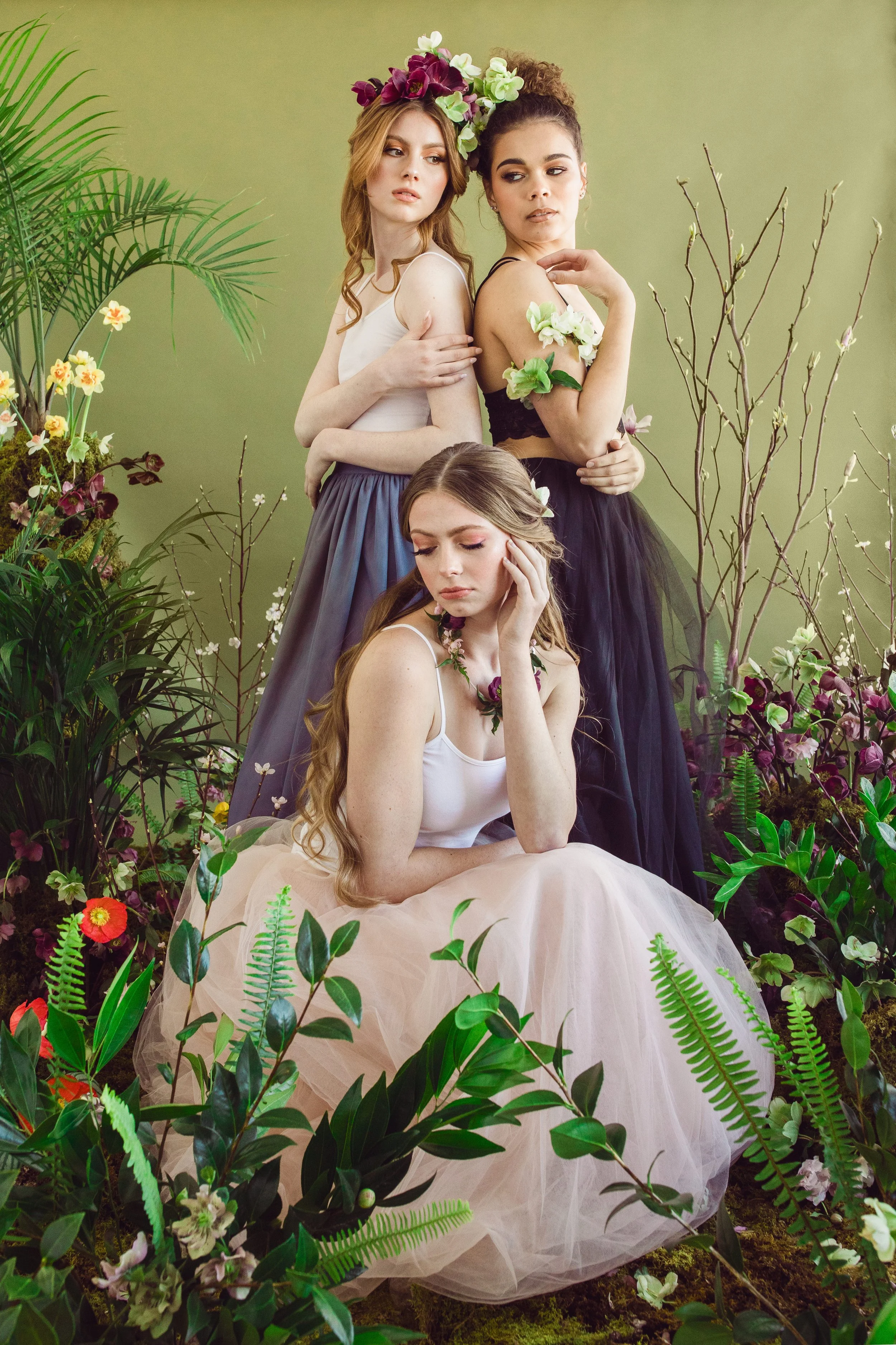 Three women in floral and tulle attire surrounded by lush greenery and flowering plants, with two standing and one sitting, posing in a studio with a green backdrop.