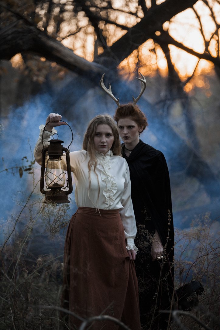 Two women dressed in vintage and fantasy costumes stand in a wooded area at sunset, one holding an old lantern, the other wearing antlers, with bluish smoke in the background.