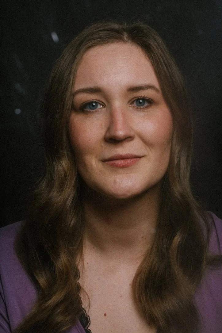 A woman with long wavy brown hair and blue eyes, wearing a purple top, looking directly at the camera with a slight smile, against a dark background.