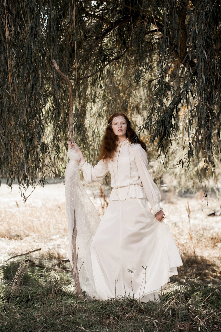 A woman with long brown hair wearing a vintage white dress stands under tree branches in a natural outdoor setting.