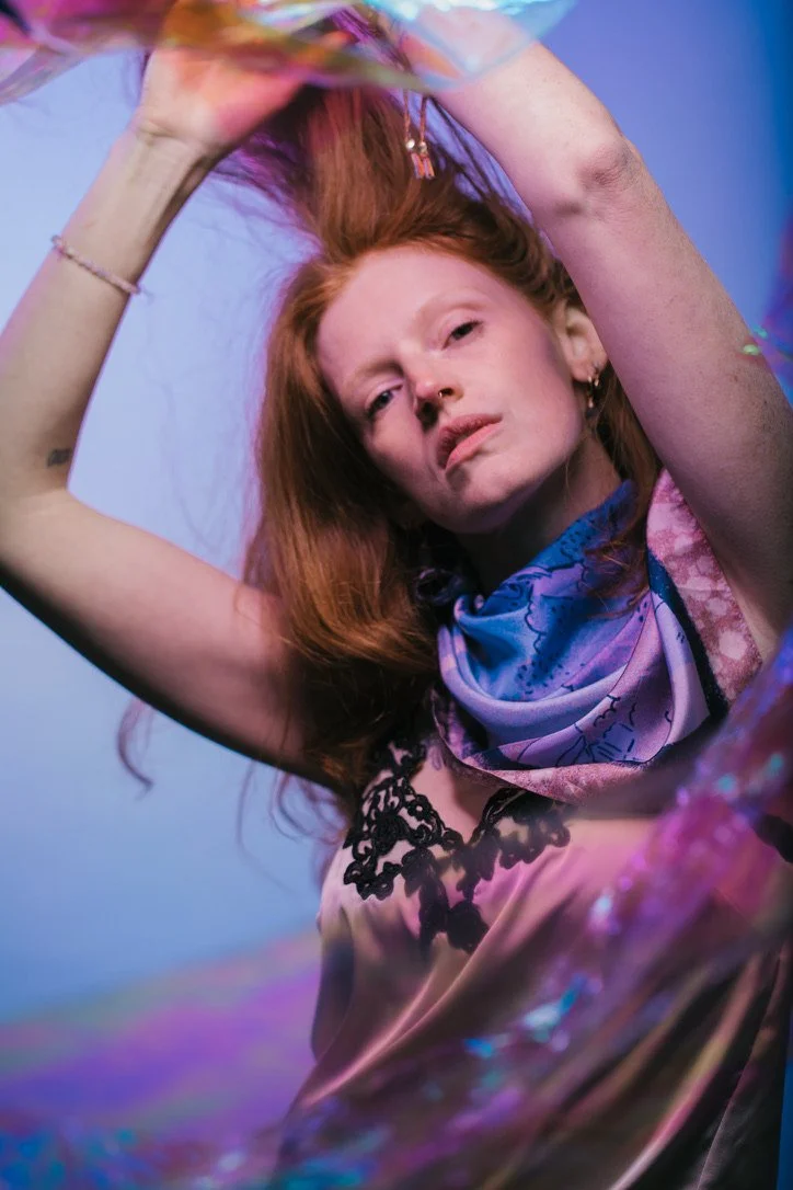 A red-haired woman with a serious expression lying down, holding her hair with one arm raised, wearing a colorful scarf and a satin dress with black lace details, against a purple-blue background.