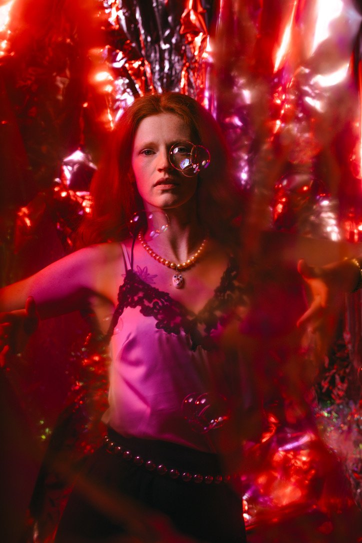 A woman with red hair and fair skin, wearing a silk camisole with black lace details, a pearl necklace with a skull pendant, and black pants with studs, stands in front of a metallic red backdrop with colorful lights, surrounded by floating soap bubb