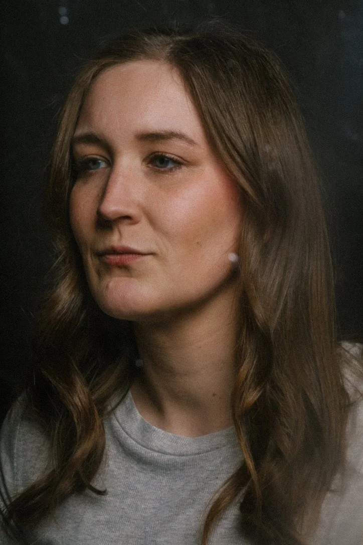 A woman with long, wavy brown hair and fair skin, wearing a gray T-shirt, looking slightly to the side against a dark background.