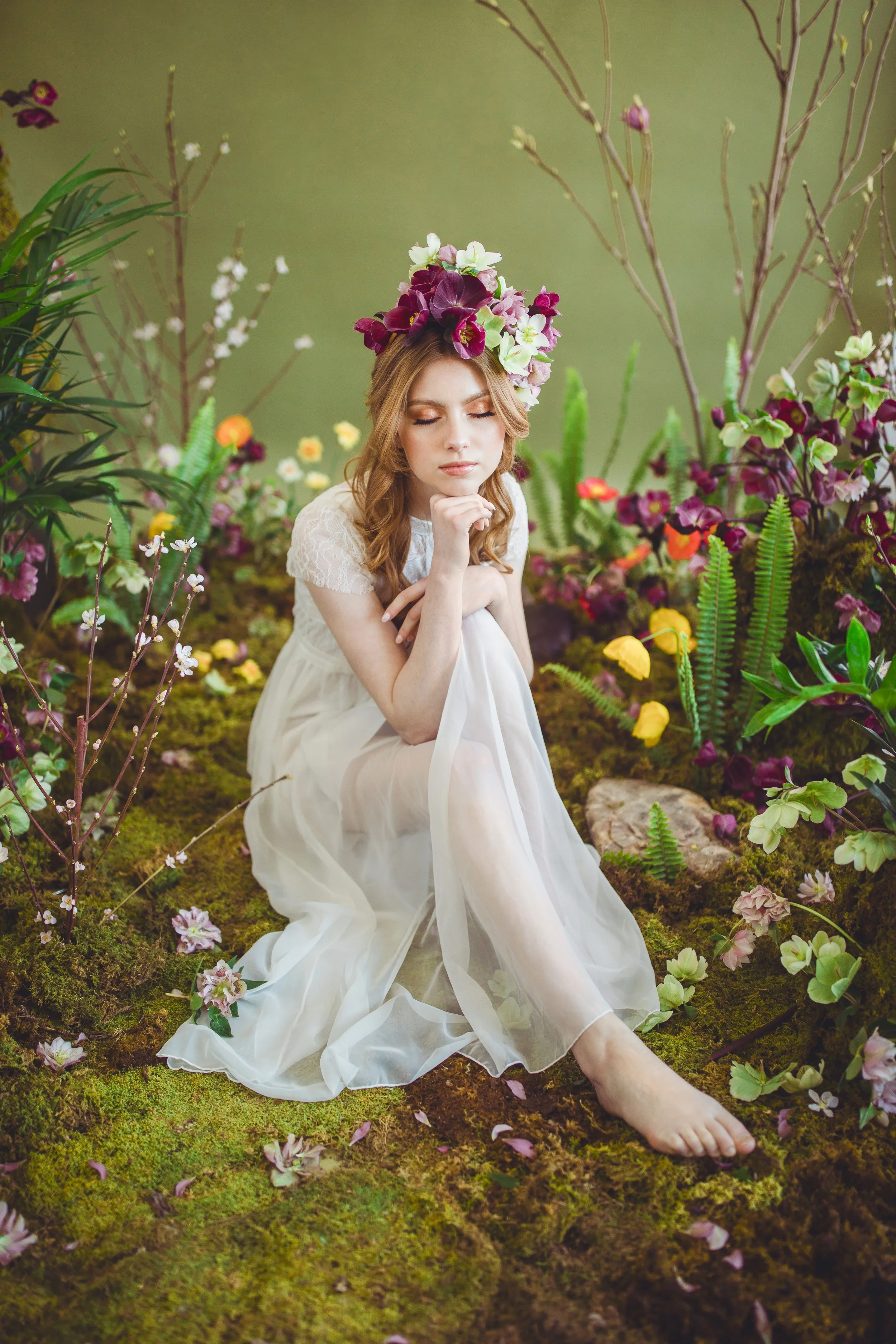 Young woman in a white dress and a floral crown sitting among lush greenery and colorful flowers.