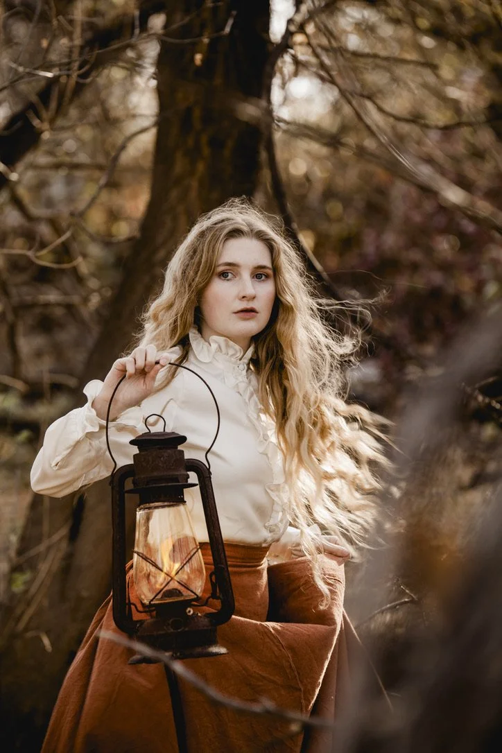 A young woman with long, wavy blonde hair, wearing a white ruffled blouse and a brown skirt, stands outdoors among trees, holding a vintage lantern with a lit flame.