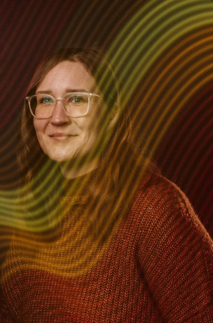 Portrait of a woman with glasses and long red hair, wearing a red knitted sweater, with yellow and red light streaks in the background.