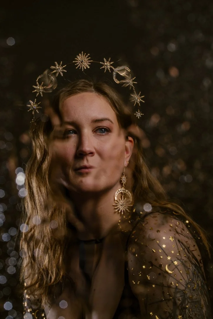 Woman wearing celestial-themed headpiece and earrings with star motifs, gazing thoughtfully, with a blurred, glittery background.