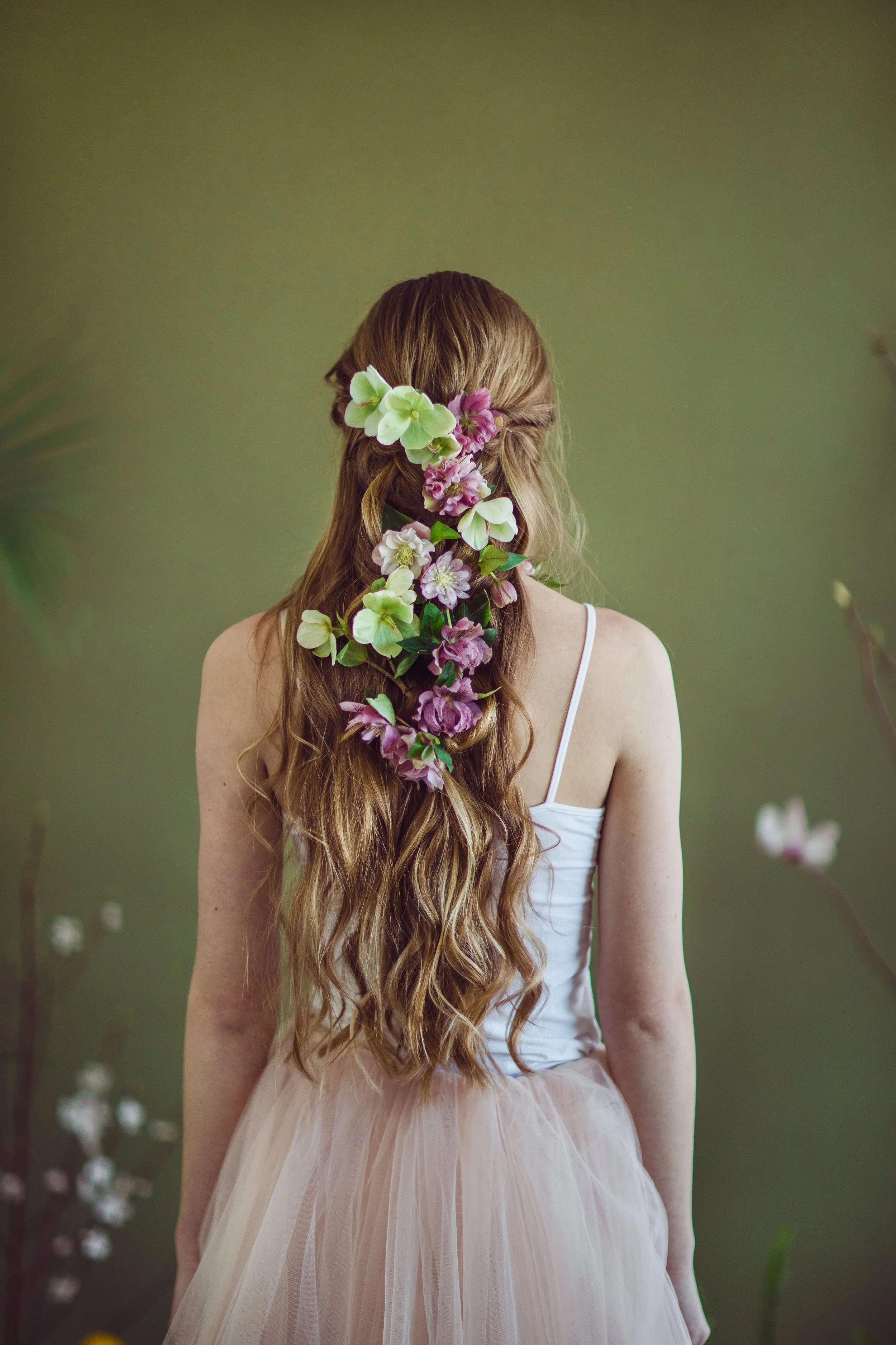 A woman with long, wavy red hair styled with a floral hairpiece of pink and white flowers, wearing a white spaghetti strap top and a light pink tulle skirt, standing against a green background.