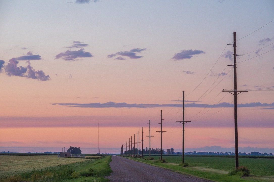 A rural landscape with a gravel road, utility poles stretching into the horizon, and open fields on each side, under a sky with scattered clouds at sunset.