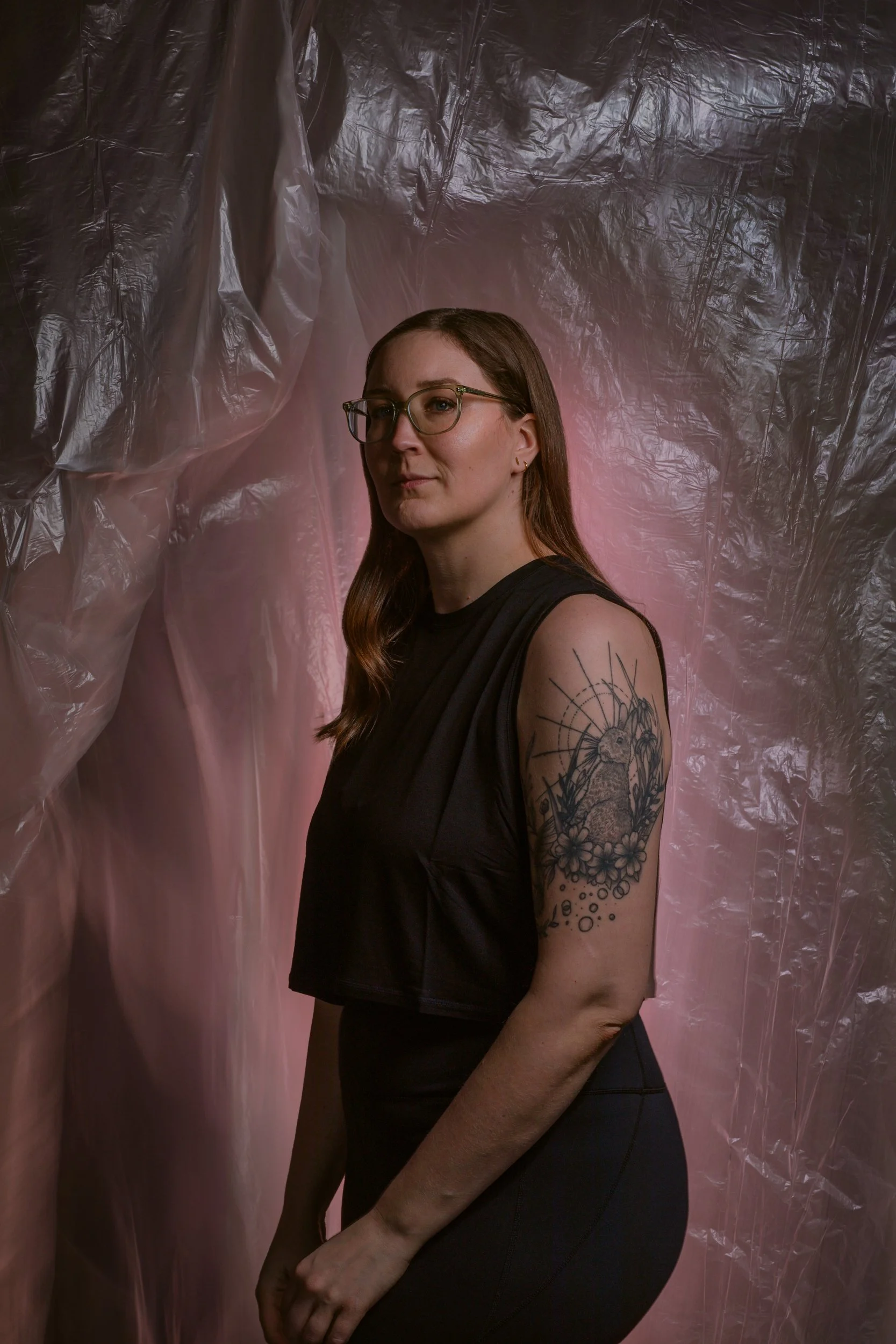 A woman with red hair, glasses, and a tattoo on her upper arm stands against a background of pink and gray plastic sheets.