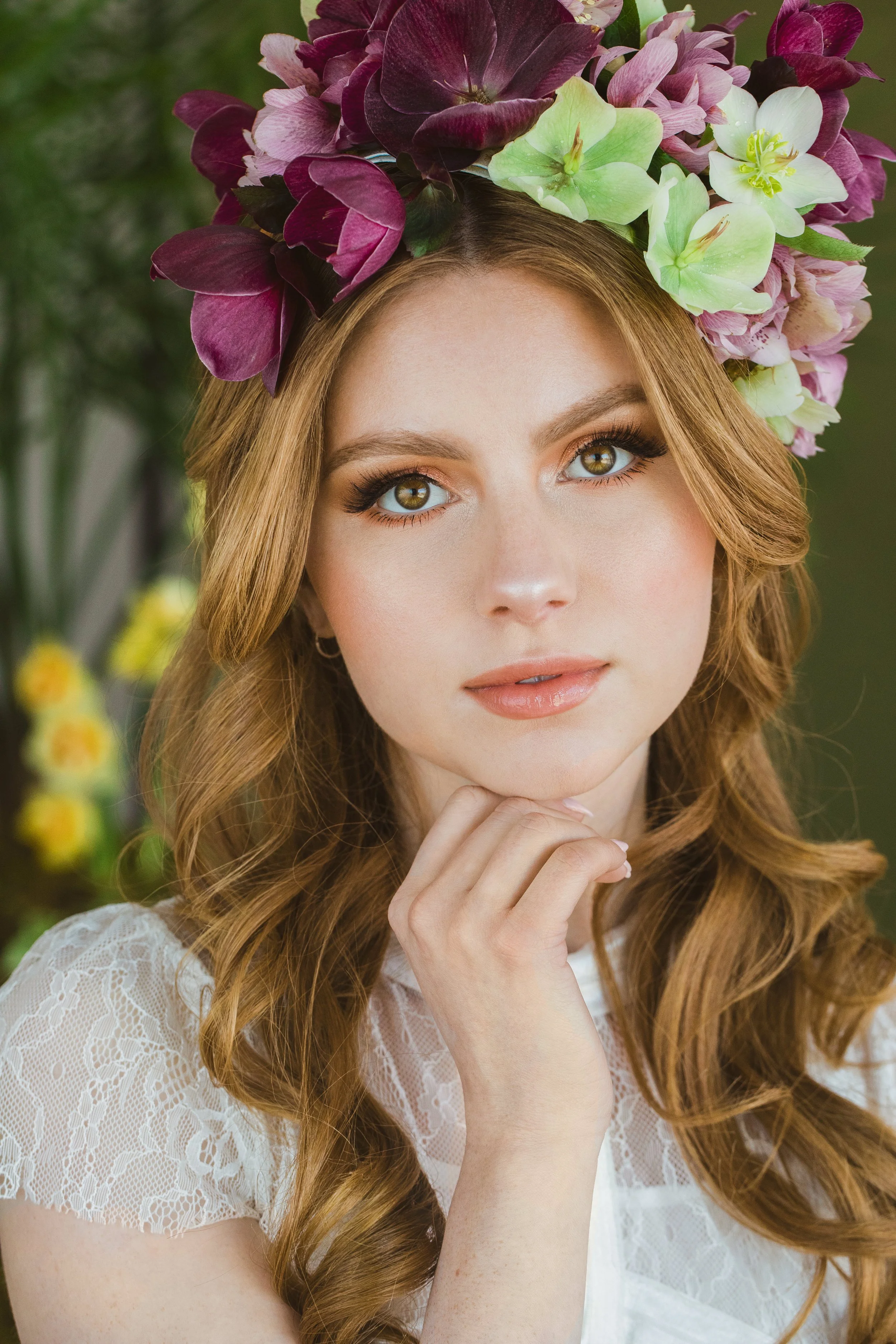 A young woman with long, wavy red hair wearing a flower crown made of purple, pink, and white flowers, and a white lace top, outdoors with a blurred green background.