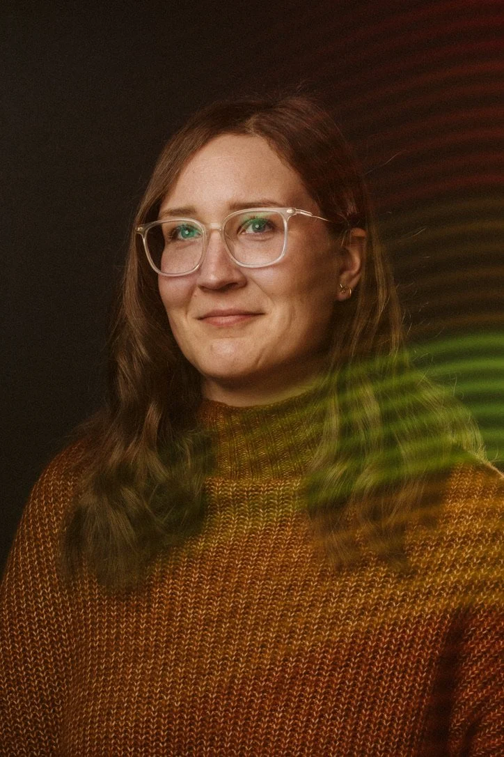 Portrait of a person with glasses and brown hair, wearing a brown knit sweater with colorful light reflections.