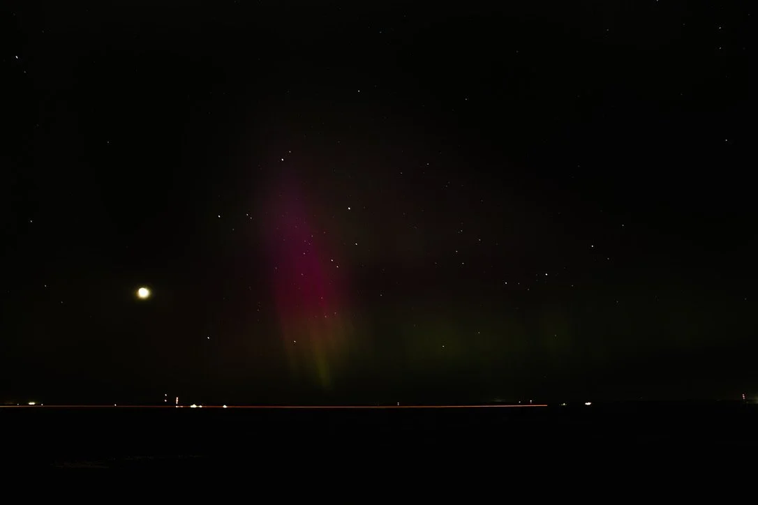 Night sky with stars, a bright moon, and faint colorful northern lights over a dark landscape.