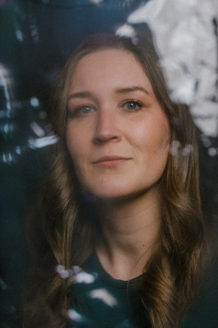 A young woman with blue eyes and long brown hair looking at the camera, with a blurred background.
