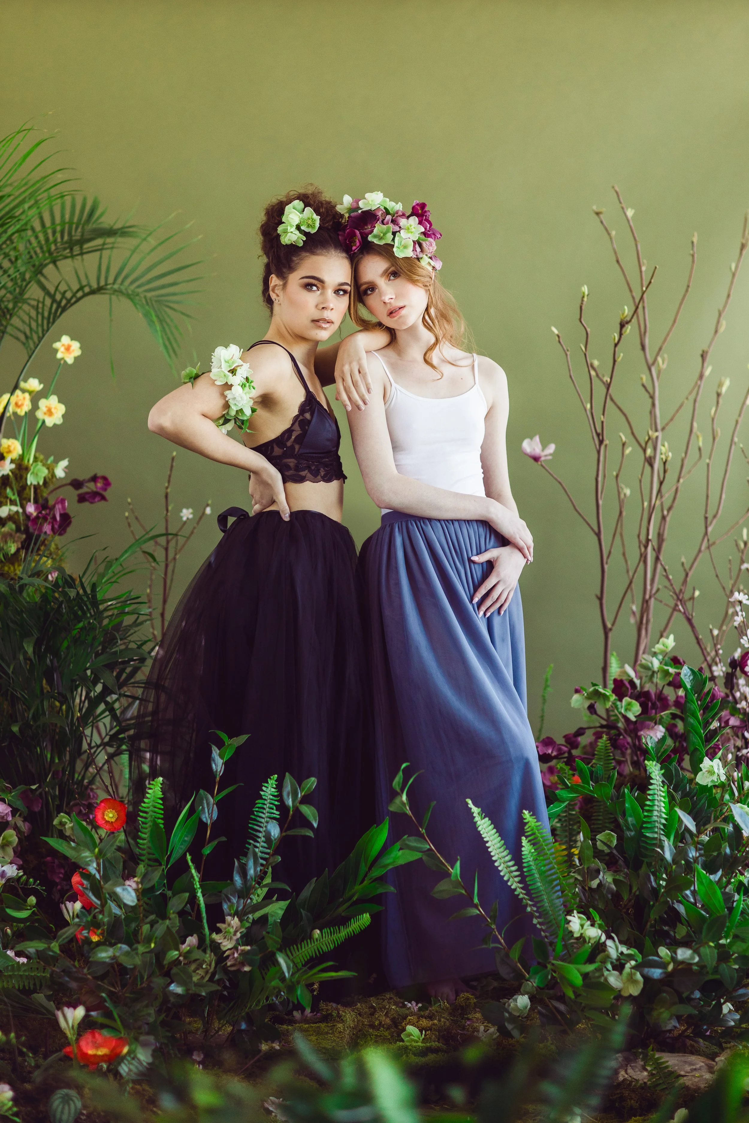 Two women wearing floral crowns and long skirts, standing among plants and flowers against a green background.
