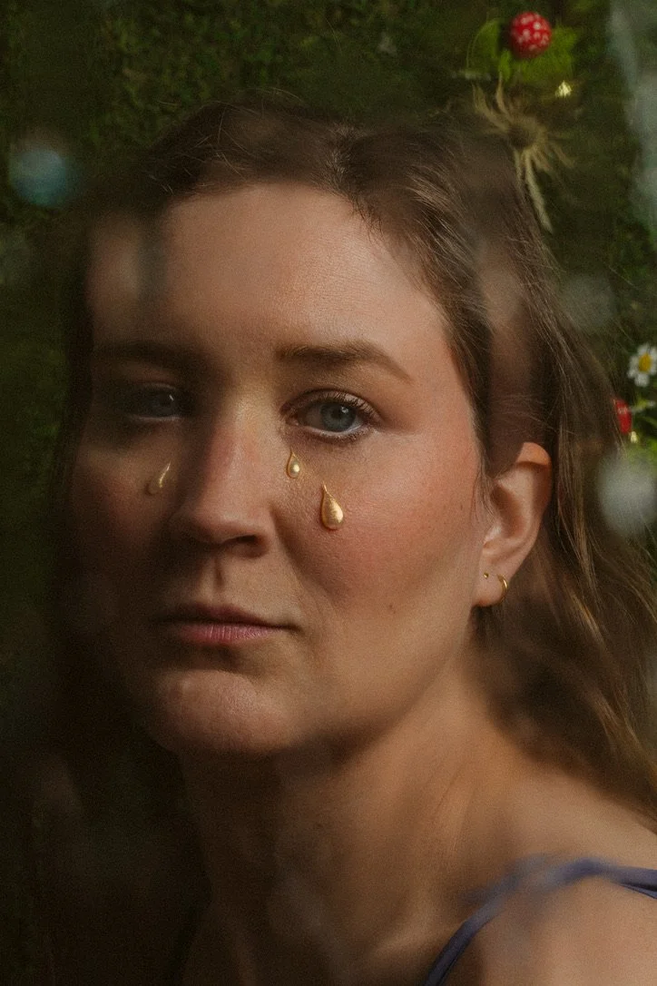 A woman with blue eyes and wavy brown hair lying on her side among green foliage with small red and white flowers scattered around.