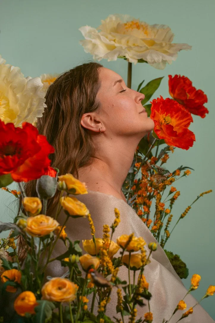 A woman with long brown hair and closed eyes surrounded by colorful flowers, including yellow, white, and red blooms, with a calm expression on her face.