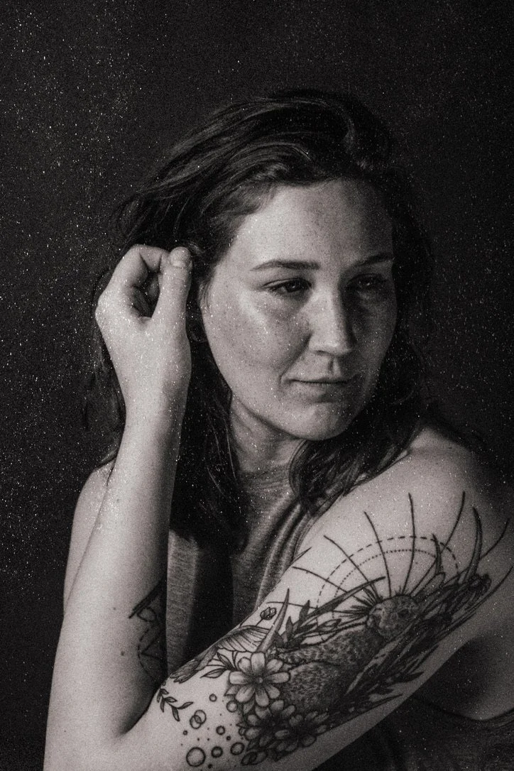 Black and white portrait of a woman with shoulder-length hair, looking contemplative, with a floral and sunburst tattoo on her upper arm, against a dark speckled background.