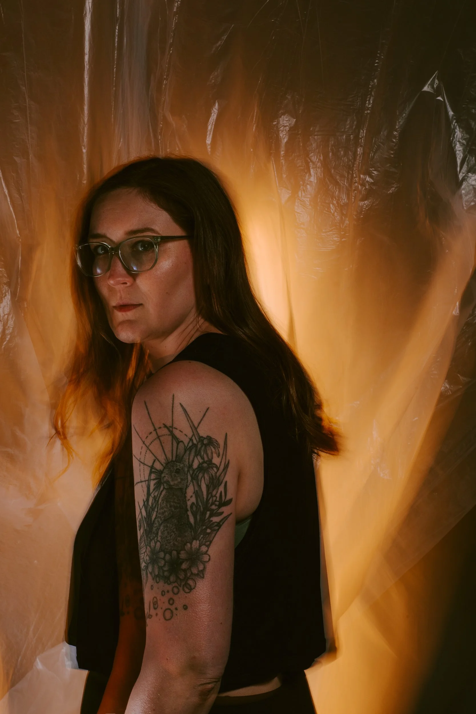 A woman with glasses and long dark hair, wearing a sleeveless black top, showing a tattoo of a rabbit surrounded by flowers and leaves on her upper arm, standing against an orange and black plastic backdrop.