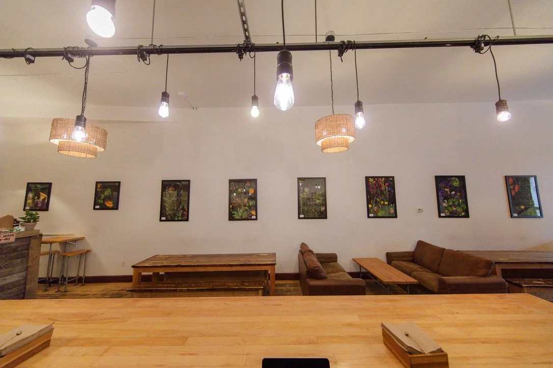 Cozy cafe or lounge with wooden tables, brown sofas, framed floral artwork on a white wall, and hanging Edison-style light bulbs.