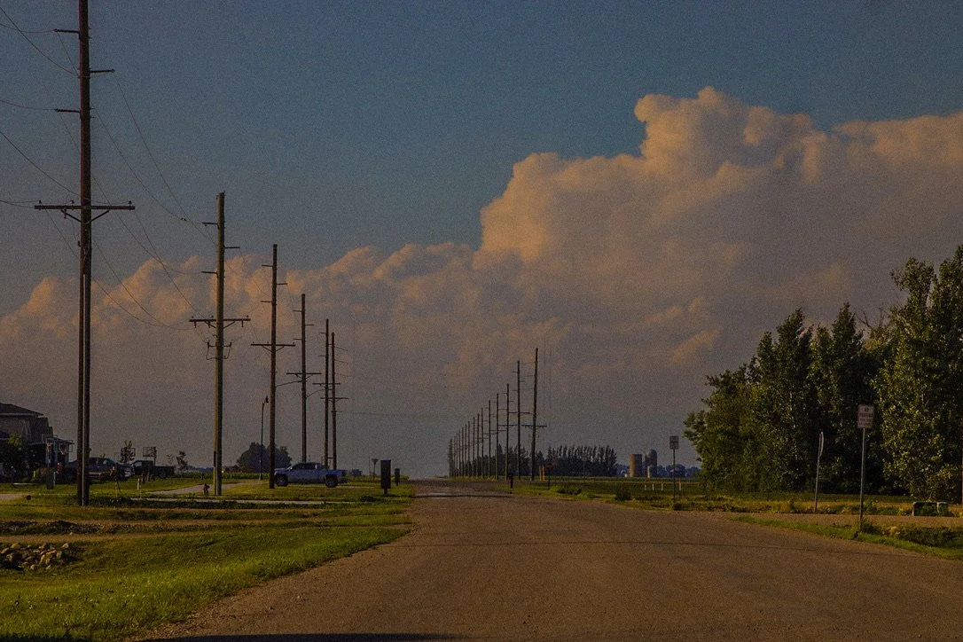 A rural street with wooden utility poles extending into the distance, green trees on the right, parked cars on the left, and a dramatic cloudy sky overhead.