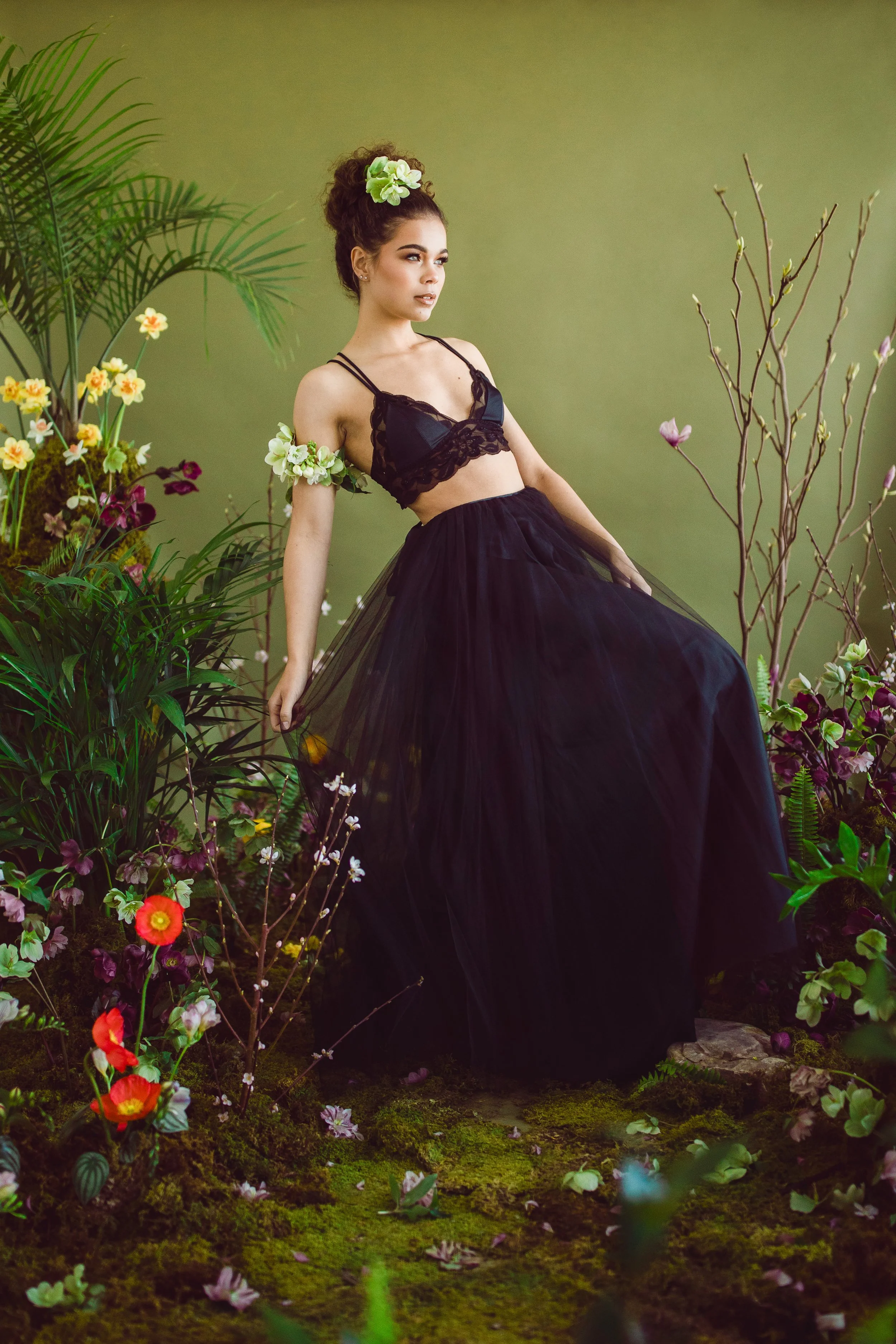 A woman dressed in a black lace bralette and long black tulle skirt posing among lush green plants and colorful flowers against a green background.