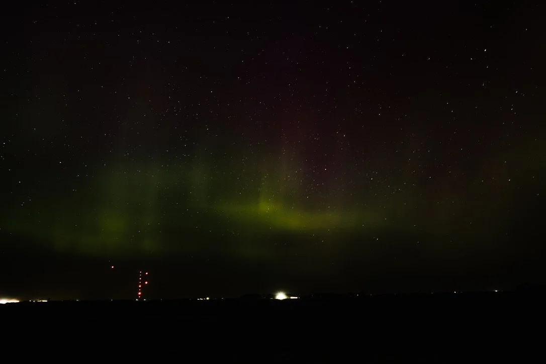 Night sky with visible stars and faint green aurora borealis above a dark landscape with distant lights.