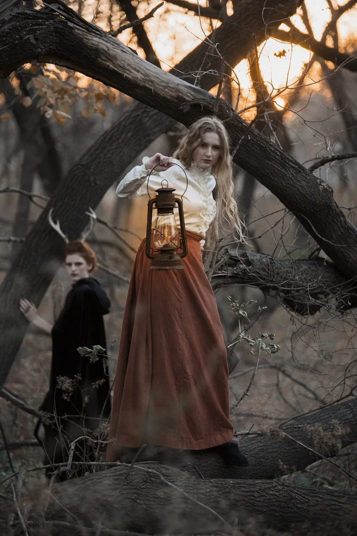 Two women in vintage clothing in a wooded area during sunset; one woman with long curly hair holds a lantern, and the other woman with red hair in a black outfit stands behind her among fallen trees.
