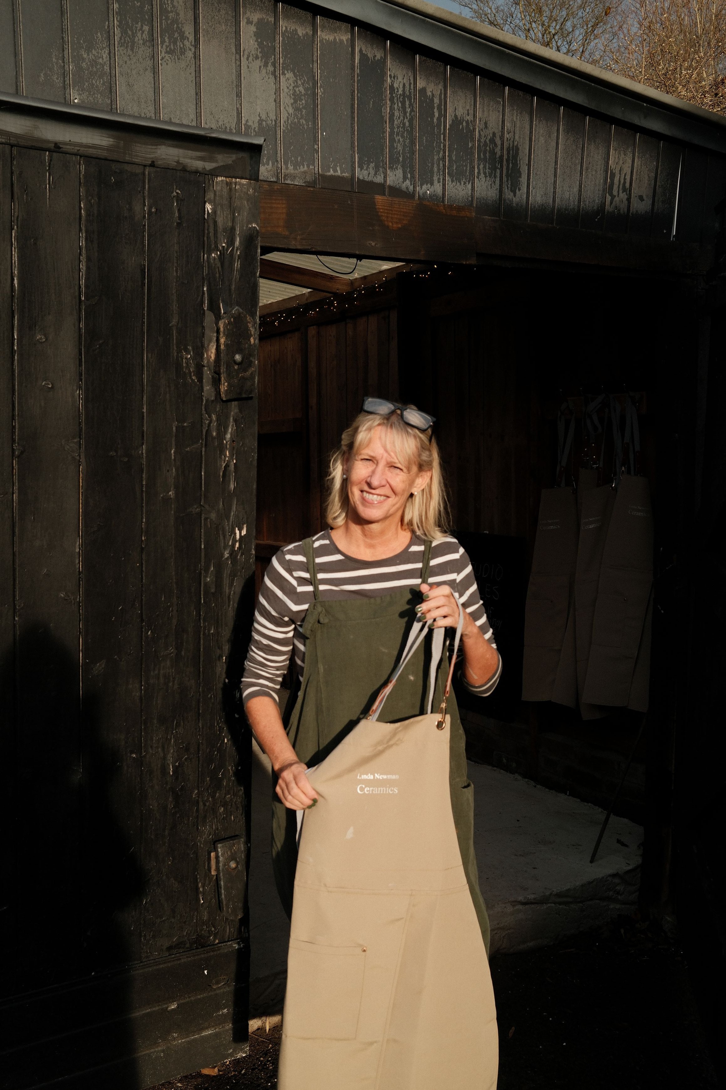A woman with blonde hair and glasses on her head is standing outside in front of a black wooden building, smiling and holding a paper bag that reads 'Linda Newman Ceramics.' She is wearing a striped shirt under an apron and holding a tote bag in her right hand.
