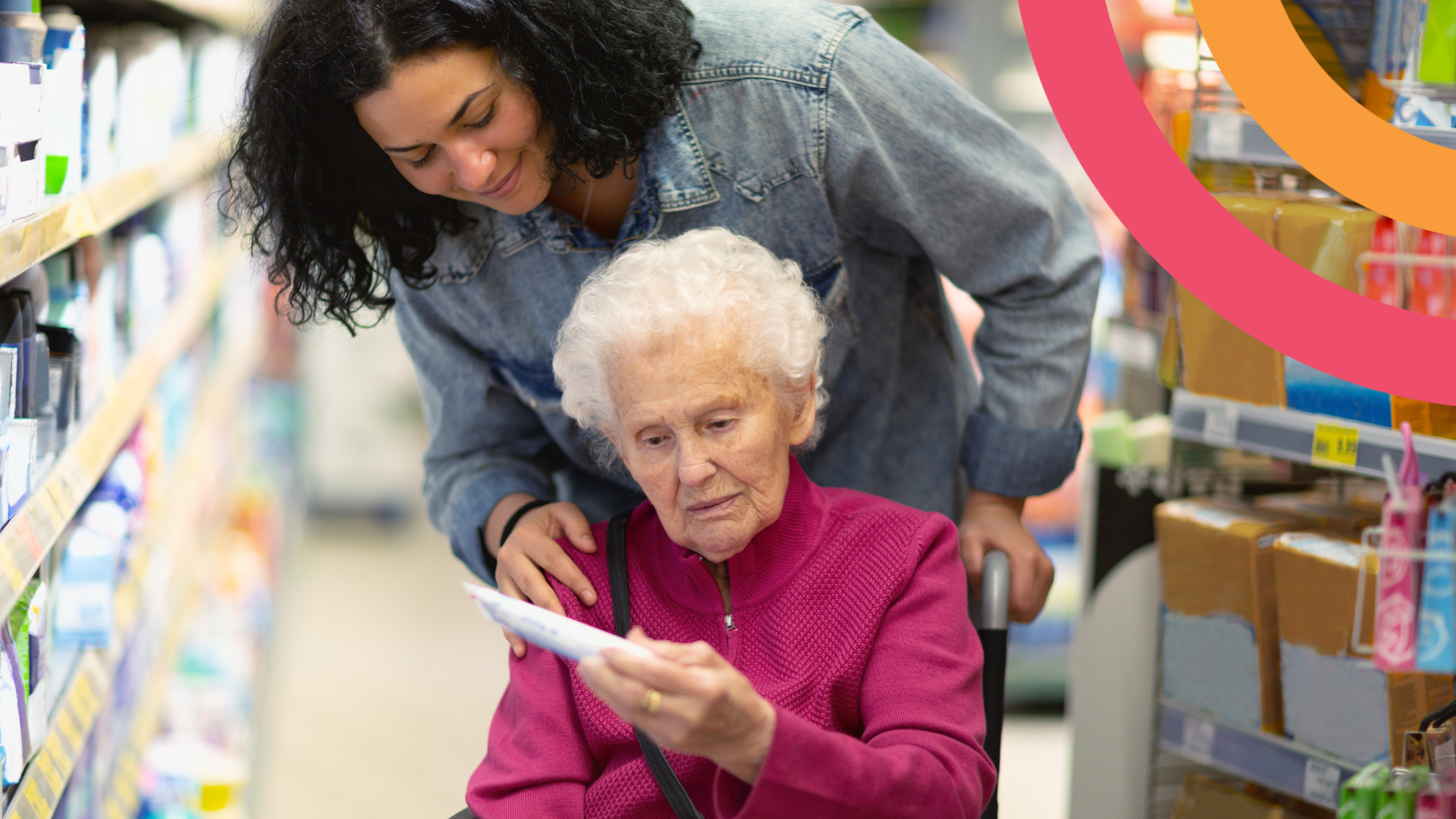 Two women are in a grocery store aisle. One is in a wheelchair reading a piece of paper. Another stands behind her with her hand on her shoulder smiling.