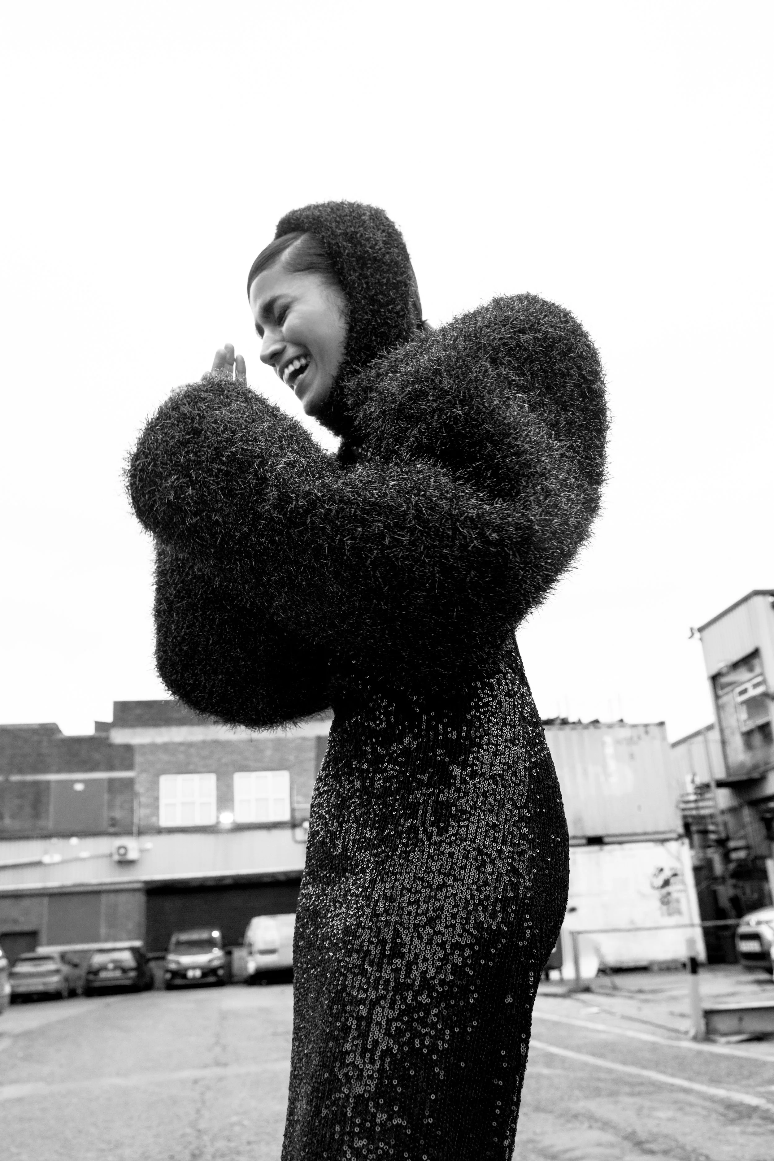 Black and white photo of a woman with short hair wearing a sequined dress and a fluffy coat, standing outdoors with buildings and parked cars in the background, smiling and posing with her hands clasped near her face.